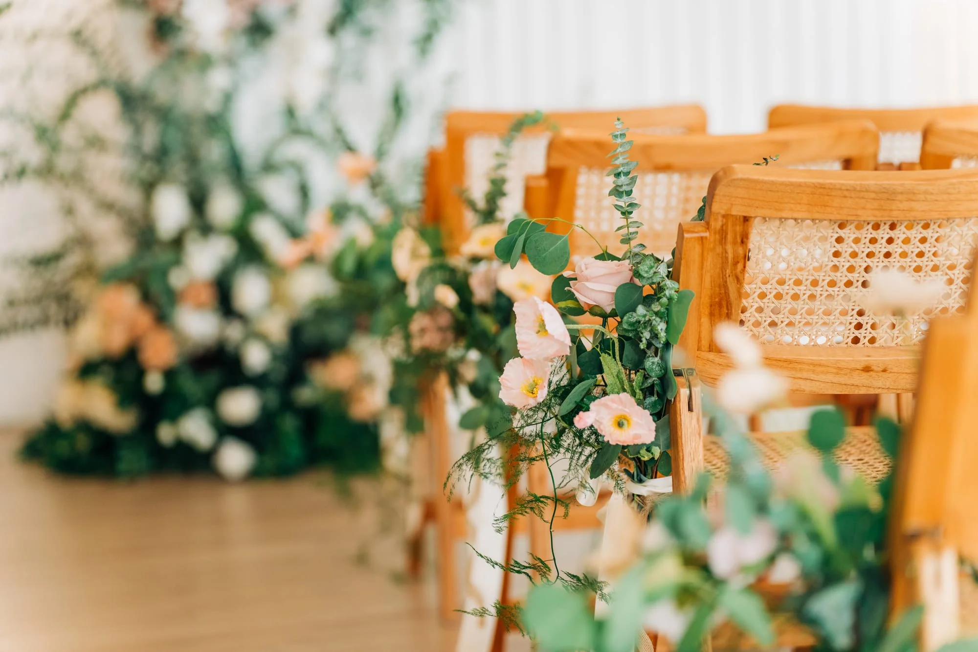 Close-up of pink poppies and eucalyptus leaves adorning a rattan ceremony chair, with a soft, dreamy bokeh background